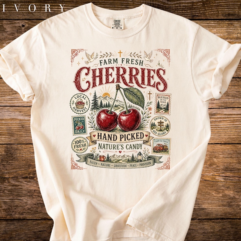 May include: Ivory-colored t-shirt featuring a vintage-style graphic with the words "Farm Fresh Cherries." The design includes two cherries, leaves, and various stamps with text like "Hand Picked" and "Nature's Candy."