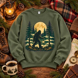May include: An olive green crewneck sweatshirt featuring a graphic illustration of a dark silhouette of Bigfoot walking through a forest of pine trees under a large, bright yellow full moon. The design includes clouds and a lantern held by Bigfoot.