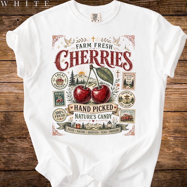 May include: White t-shirt featuring a graphic design with the words "Farm Fresh Cherries" in a vintage style. The design includes illustrations of cherries, leaves, and other elements. The shirt also has additional text and illustrations.