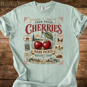 May include: Light blue t-shirt featuring a graphic of two cherries with the text "Farm Fresh Cherries". The design includes illustrations of a mountain scene, birds, and other decorative elements. The shirt is made of a soft material.