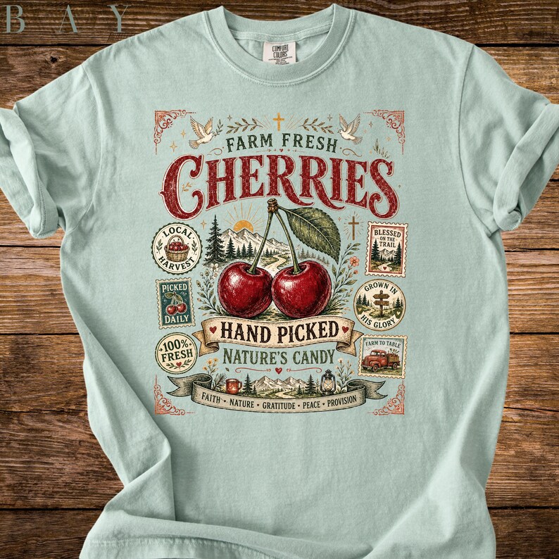 May include: Light blue t-shirt featuring a graphic of two cherries with the text "Farm Fresh Cherries". The design includes illustrations of a mountain scene, birds, and other decorative elements. The shirt is made of a soft material.