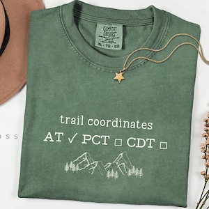 May include: A green Comfort Colors t-shirt with the text "trail coordinates" and check boxes for AT, PCT, and CDT. A gold star necklace rests on the shirt. The shirt also features a mountain and tree graphic.