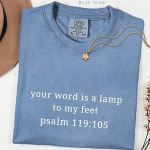 Puede incluir: Una camiseta azul vaquero con el texto "your word is a lamp to my feet psalm 119:105" en blanco. Un collar con un colgante de estrella dorada descansa sobre la camisa.