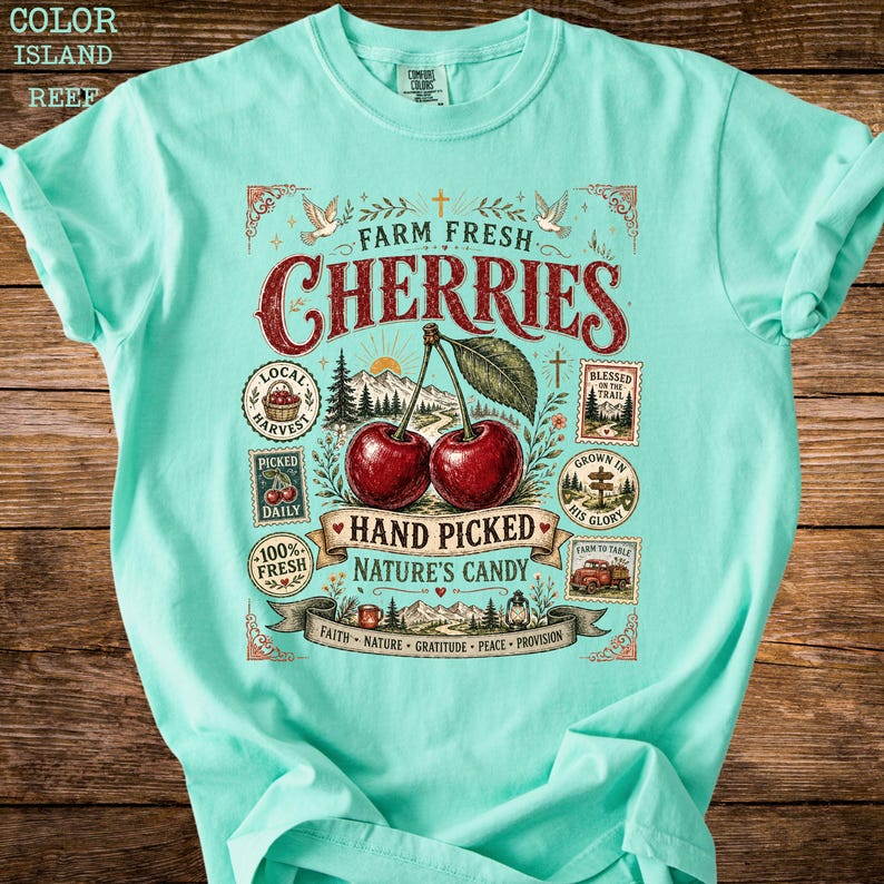 May include: A light turquoise t-shirt with a graphic featuring two cherries and the words "Farm Fresh Cherries." The design includes illustrations of a mountain scene, doves, and text that reads "Hand Picked Nature's Candy."