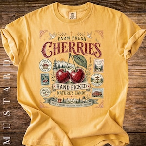 May include: Mustard-colored t-shirt featuring a vintage-style graphic with the words "Farm Fresh Cherries." The design includes two cherries, text, and decorative elements. The shirt is made of a soft material.