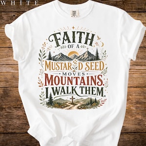 Könnte beinhalten: Weißes T-Shirt mit einem grafischen Design. Das Design zeigt den Text "Faith of a Mustard Seed Moves Mountains I Walk Them" mit Bergen, einem Kreuz und botanischen Elementen in Grün, Braun und Gold.