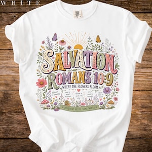 Puede incluir: Camiseta blanca con un diseño floral colorido y el texto "SALVATION ROMANS 10:9 WHERE THE FLOWERS BLOOM". El diseño incluye mariposas, flores y la frase "DIFFERENT NEEDS. SAME WORTH".