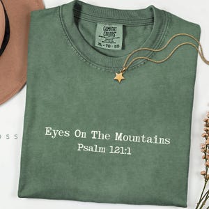 May include: A sage green t-shirt with the text "Eyes On The Mountains Psalm 121:1" in white. A gold star pendant necklace rests on the shirt. The shirt has a Comfort Colors tag.
