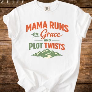 Könnte beinhalten: Weißes T-Shirt mit dem Text "MAMA RUNS on Grace AND PLOT TWISTS" in Orange und Grün. Das Design beinhaltet eine Berglandschaft. Das Shirt ist aus weichem Material.