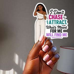 May include: A sticker featuring a stylish illustration of a woman in a white jumpsuit, with the text "I Don't CHASE - I ATTRACT. What's Meant FOR ME WILL FIND ME." The sticker is held by a hand with manicured nails.