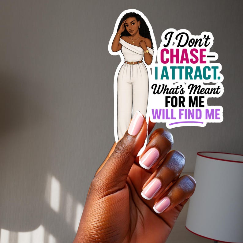 May include: A sticker featuring a stylish illustration of a woman in a white jumpsuit, with the text "I Don't CHASE - I ATTRACT. What's Meant FOR ME WILL FIND ME." The sticker is held by a hand with manicured nails.