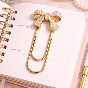 May include: A gold-colored paper clip with a bow-shaped top, encrusted with small, clear crystals. The clip is resting on a pink and white notebook, with the word "MONDAY" printed on the page.