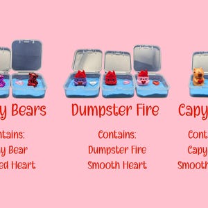 May include: Three small, open plastic containers with blue sand and miniature figurines. The containers are labeled "Shiny Bears," "Dumpster Fire," and "Capybara." Each contains a small figurine and a heart. The background is a light pink.