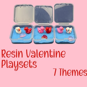 May include: Three clear plastic containers with open lids, each containing blue sand and small resin figurines. The figurines include ducks and heart-shaped objects in red, pink, and white. Text on the image reads "Resin Valentine Playsets" and "7 Themes".