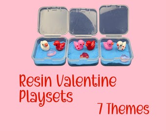 May include: Three clear plastic containers with open lids, each containing blue sand and small resin figurines. The figurines include ducks and heart-shaped objects in red, pink, and white. Text on the image reads "Resin Valentine Playsets" and "7 Themes".