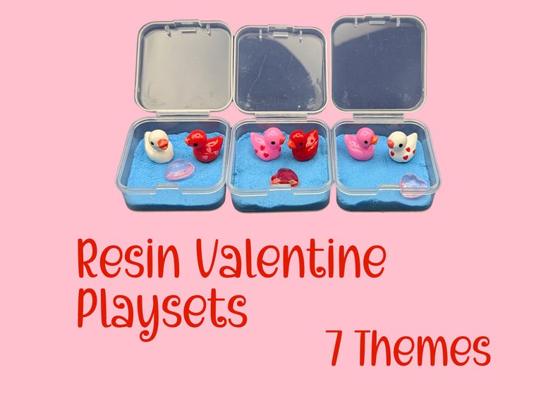 May include: Three clear plastic containers with open lids, each containing blue sand and small resin figurines. The figurines include ducks and heart-shaped objects in red, pink, and white. Text on the image reads "Resin Valentine Playsets" and "7 Themes".