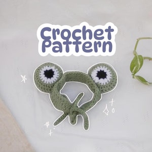 May include: Green crocheted frog headband with white and black eyes. The headband is on a white background with a small plant in the corner.