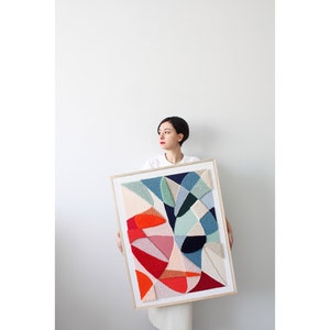 May include: A woman holds a framed abstract geometric artwork with a white background and colorful shapes in shades of red, orange, pink, blue, and green.