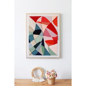 May include: A rectangular wall art piece featuring a textured, woven fiber design in a light wooden frame. The composition displays abstract, overlapping geometric shapes in shades of red, pink, teal, navy blue, beige, and light green.