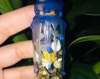 May include: A small glass bottle filled with layers of herbs, crystals, and a white powder. The bottle is sealed with a cork and blue wax that has dripped down the sides. The background is dark green foliage.