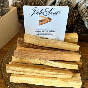 May include: A stack of Palo Santo sticks on a decorative glass plate. The sticks are light brown and have a natural wood grain. The text on the card reads "Palo Santo" and "Cleanses negative energies, Invites in peace and harmony, Increases creativity, Promotes spiritual healing".