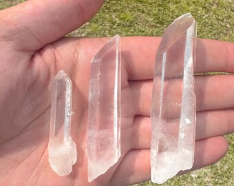 May include: Three clear quartz crystal points of varying sizes, held in a hand against a green grassy background.