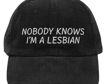 May include: Black corduroy baseball cap with white embroidered text that reads "Nobody Knows I'm A Lesbian".