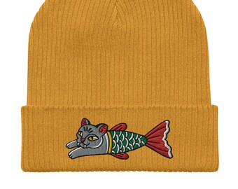 May include: A mustard yellow knitted beanie with a gray cat fish design embroidered on the front. The cat fish has green and red scales and a red tail fin.