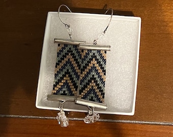 May include: A pair of silver earrings with geometric patterned beads in shades of blue, black, and gold. The earrings have a crystal drop at the bottom.