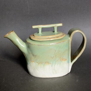 Op de afbeelding: Handgemaakte keramische theepot met een lichtgroen glazuur. De theepot heeft een afgeronde body, een gebogen handvat en een tuit. Het deksel heeft een klein handvat. De bodem van de theepot is wit.