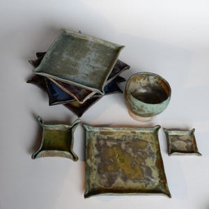Può includere: Una collezione di pezzi in ceramica fatti a mano nei toni del verde, blu e marrone. Il set comprende vassoi quadrati di varie dimensioni, una piccola ciotola e piatti quadrati con bordi curvi. I pezzi hanno un aspetto rustico e strutturato.