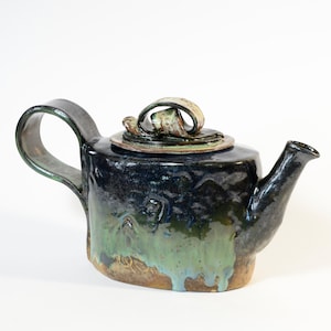 Op de afbeelding: Handgemaakte keramische theepot met een donkerblauwe, groene en bruine glazuur. De theepot heeft een unieke handgreep en een decoratief deksel met een gekruld ontwerp. Een functioneel en artistiek stuk.