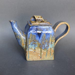 Peut inclure: Une théière en céramique faite à la main, de forme carrée et dotée d'une anse incurvée. La théière présente un glaçage bleu avec des accents bruns et beiges, créant un effet dégoulinant. Le couvercle a une petite poignée. Un objet de cuisine unique.