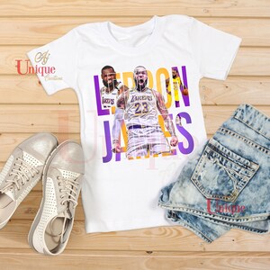 Könnte beinhalten: Weißes T-Shirt mit einer Grafik von LeBron James im Lakers-Trikot und dem Text "LEBRON JAMES" in Lila und Gold. Das Bild zeigt Jeansshorts und silberne Turnschuhe.
