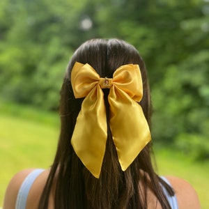 Puede incluir: Un gran lazo de satén amarillo con un dije de corona dorada. El lazo está atado en la parte posterior del cabello de una persona.