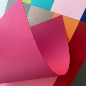 May include: A collection of colorful paper sheets in various shades of pink, red, orange, green, blue, and gray. The sheets are arranged in a staggered pattern, creating a visually appealing abstract design.