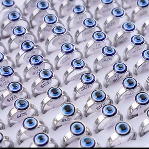May include: A collection of silver-toned rings, each featuring a blue and black evil eye design. The rings are arranged in rows, with size labels visible on some. The evil eye is a popular symbol for protection.