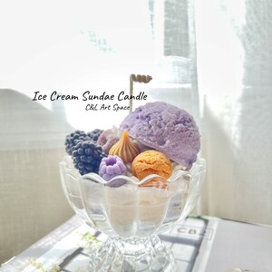 May include: A purple, white, and brown ice cream sundae candle in a clear glass dish. The candle is decorated with blackberries, raspberries, and a caramel swirl. The text "Ice Cream Sundae Candle" and "C&L Art Space" is visible on the image.