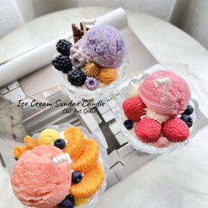 May include: Three ice cream sundae candles in glass dishes. The candles are decorated with various toppings, including fruit, chocolate, and sprinkles. The candles are labeled "Ice Cream Sundae Candle C&L Art Space".