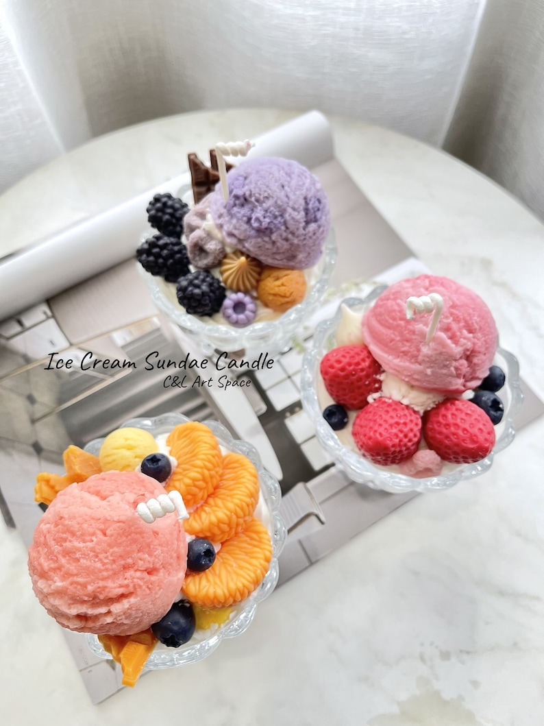 May include: Three ice cream sundae candles in glass dishes. The candles are decorated with various toppings, including fruit, chocolate, and sprinkles. The candles are labeled "Ice Cream Sundae Candle C&L Art Space".