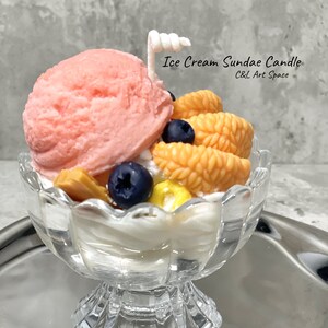 May include: A pink and white ice cream sundae candle in a clear glass dish with a white cherry on top. The candle is decorated with orange and yellow fruit slices and blueberries. The text "Ice Cream Sundae Candle" and "C&L Art Space" is printed on the image.