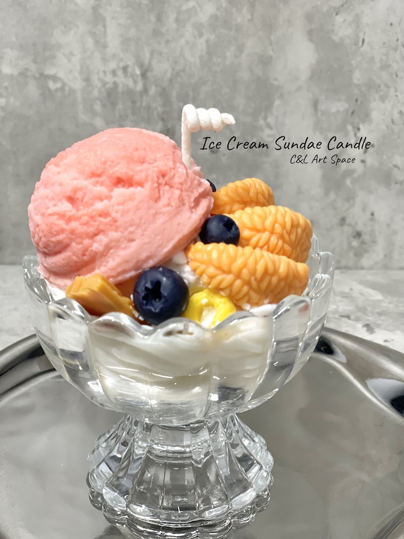 May include: A pink and white ice cream sundae candle in a clear glass dish with a white cherry on top. The candle is decorated with orange and yellow fruit slices and blueberries. The text "Ice Cream Sundae Candle" and "C&L Art Space" is printed on the image.