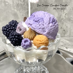 May include: A purple and white ice cream sundae candle in a clear glass dish with a white chocolate stick on top. The candle is decorated with blackberries, a chocolate bar, and a white chocolate swirl. The candle is on a silver tray. The text "Ice Cream Sundae Candle" and "C&L Art Space" is on the image.