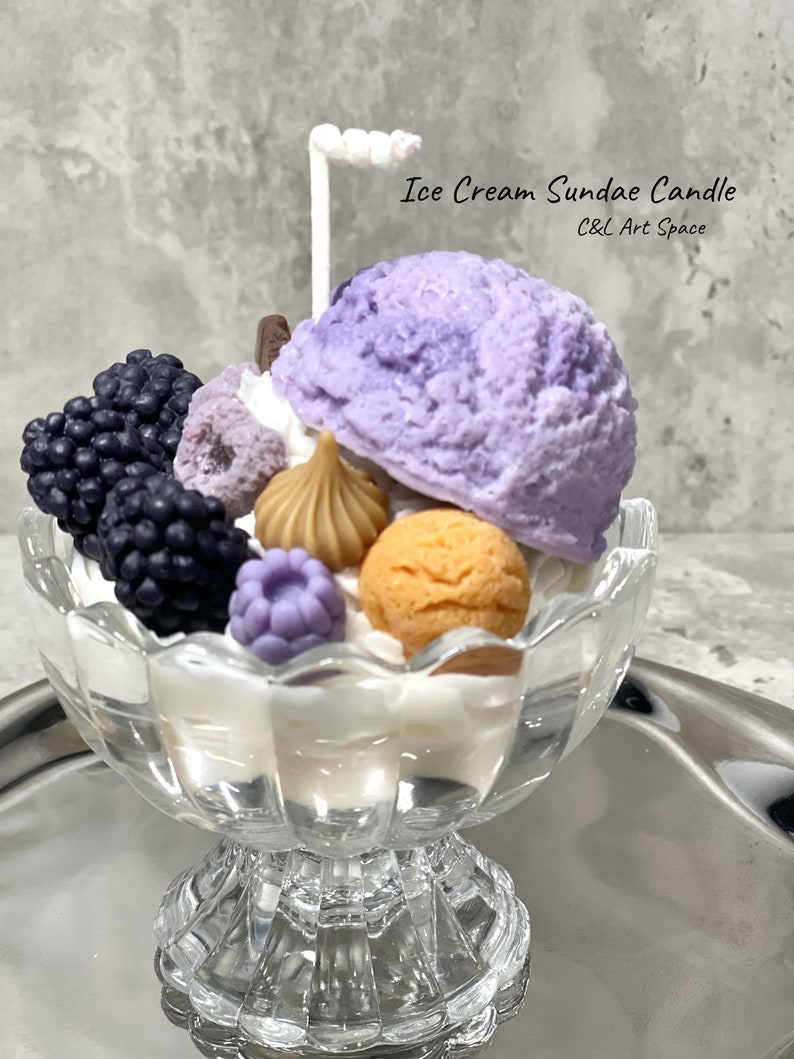 May include: A purple and white ice cream sundae candle in a clear glass dish with a white chocolate stick on top. The candle is decorated with blackberries, a chocolate bar, and a white chocolate swirl. The candle is on a silver tray. The text "Ice Cream Sundae Candle" and "C&L Art Space" is on the image.