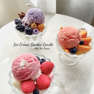 May include: Three ice cream sundae candles in glass dishes. The candles are decorated with various toppings, including fruit, chocolate, and sprinkles. The candles are labeled "Ice Cream Sundae Candle C&L Art Space".