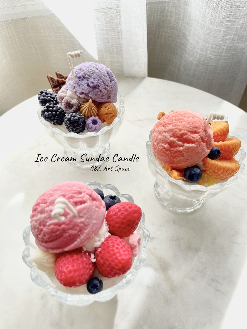 May include: Three ice cream sundae candles in glass dishes. The candles are decorated with various toppings, including fruit, chocolate, and sprinkles. The candles are labeled "Ice Cream Sundae Candle C&L Art Space".