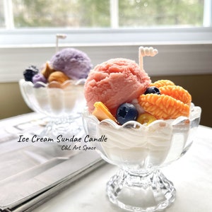 May include: Two glass ice cream sundae candle holders with a pink and purple ice cream candle in each. The candles are topped with a white chocolate swirl and blueberries. The text "Ice Cream Sundae Candle C&C Art Space" is printed below the candles.