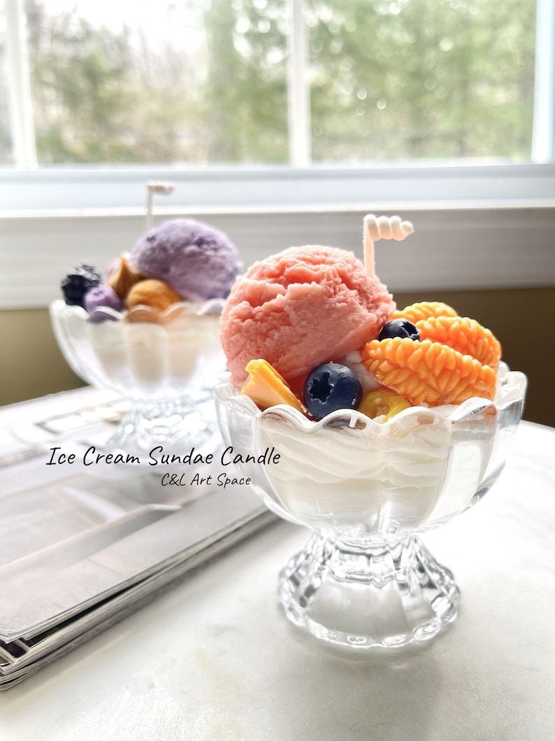 May include: Two glass ice cream sundae candle holders with a pink and purple ice cream candle in each. The candles are topped with a white chocolate swirl and blueberries. The text "Ice Cream Sundae Candle C&C Art Space" is printed below the candles.