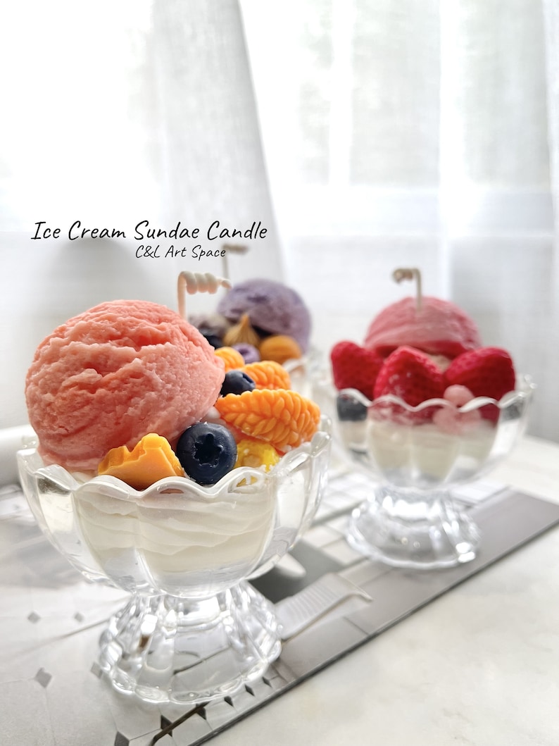 May include: Three ice cream sundae candles in glass dishes. The candles are decorated with pink, white, and red ice cream, yellow and orange fruit, and blue blueberries. The candles are labeled "Ice Cream Sundae Candle" and "C&L Art Space".
