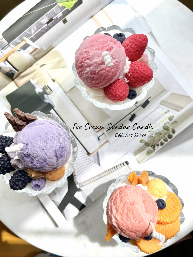 May include: Three ice cream sundae candles in clear glass dishes. The candles are decorated with various toppings, including strawberries, blueberries, chocolate, and orange slices. The candles are labeled "Ice Cream Sundae Candle" and "C&L Art Space".